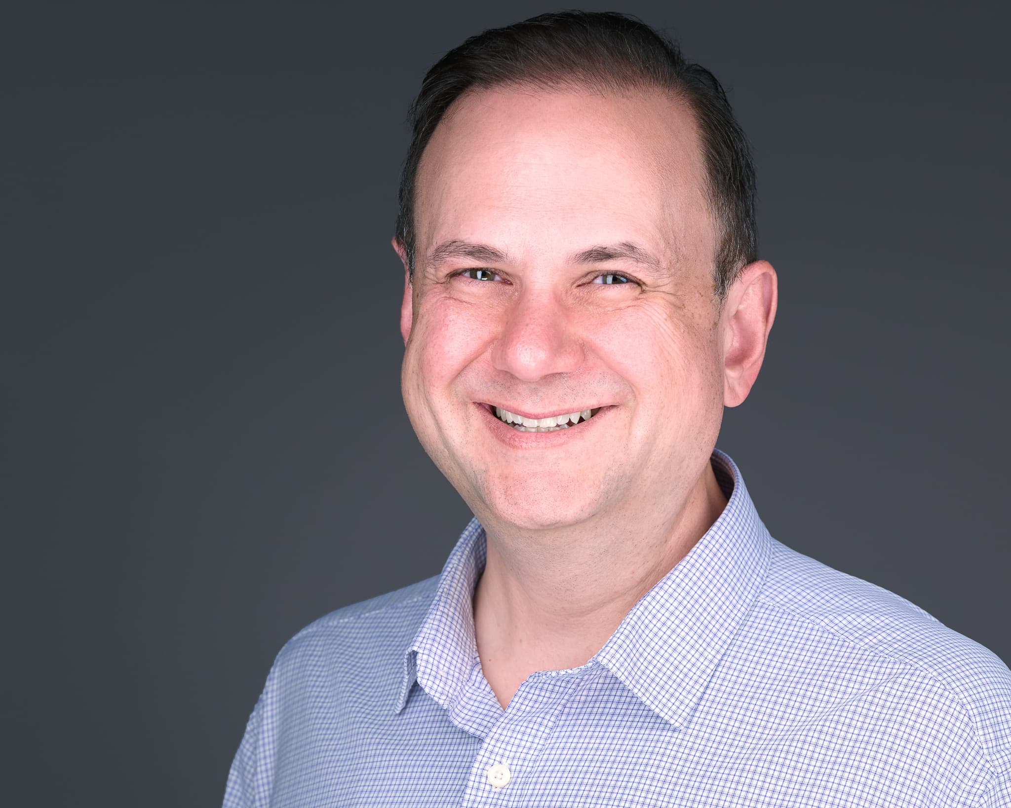 Chris Kiklas in a light blue checked shirt, smiling against a gray studio backdrop.